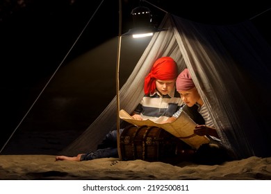 Children Dressed As Sailors Play In A Tent On The Sand At Night By The Light Of A Lantern And Pretend To Be Pirates Looking For Treasure On A Map. Funny Kids Dream Of Adventures And Travels.