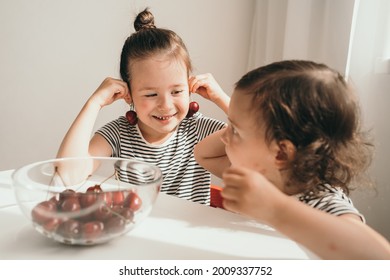 Children Eat Delicious And Juicy Cherries. A Small Child Indulges In Food. The Girl Puts The Cherry To Her Ear Like An Earring. Two Sisters Cheerfully Eat Summer Berries