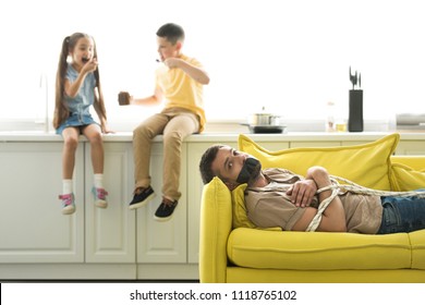 Children Eating Chocolate And Father Lying Tied On Sofa At Home