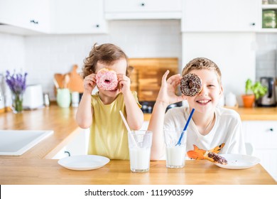 Children Eating Donuts On The White Kitchen At Home. Child Is Having Fun With Donuts. Tasty Food For Kids.