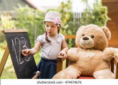 Children Education Concept. Cute Toddler Playing Teacher Role Game With Her Teddy Bear Friend Outdoors. Happy Kid Leaning Numbers