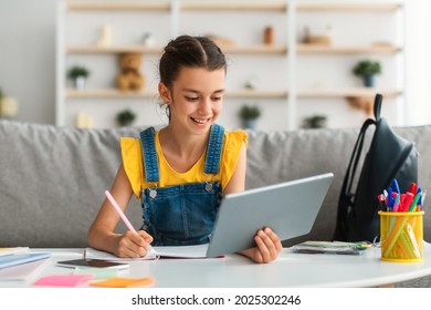Children and Education Concept. Portrait of positive teen girl using doing homework writing in notebook sitting on couch at desk, holding and using tablet pad computer, enjoying distance homeschooling - Powered by Shutterstock