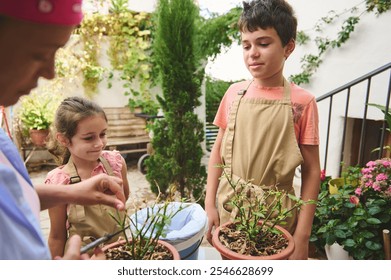 Children engaged in a gardening activity, learning from an adult in a sunlit backyard. They are wearing aprons and handling potted plants, surrounded by lush greenery and flowers. - Powered by Shutterstock