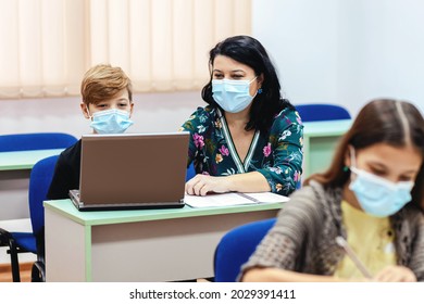 Children With Face Mask Back To School In Classroom After Covid-19 Lockdown - Teacher Helping Kid To Use Computer Doing Hybrid Education Class After School Reopening