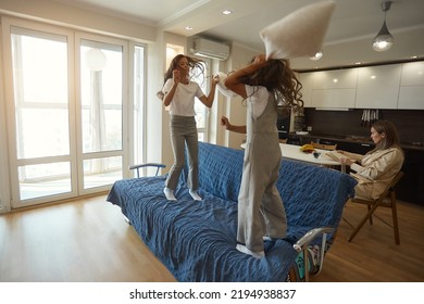 Children Fight With Pillows On The Sofa, Two Sisters Play On The Sofa With Pillows, Run Around The Apartment. Mom Keeps Calm. Mother Of The Year