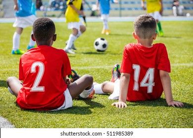 Children Football Soccer Tournament. Kids Playing Football Match. Two Boys Friends Caucasian And African In Red Shirts Watching Soccer Game Competition. 