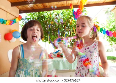 Children Girls Having Fun At A Birthday Party In A Home Garden Blowing Bubbles Joyfully, House Exterior. Kids Playing With Decorations And Balloons, Outdoors. Fun And Activities On Holiday, Sunny Day.