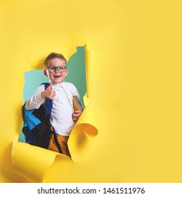 Children Go Back To School. Pupil Of Primary School With Backpack And Book Breaking Yellow Paper Wall. Beginning Of Lessons. First Day Of Fall. Elementary Student.