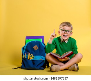 Children Go Back To School. Stylish Boy Doing Homework At Home With Backpack Full Of Books, Pencils. Pupil Reading A Book, Writing And Painting. Kid Is Drawing. Child In Glasses On Yellow Wall.