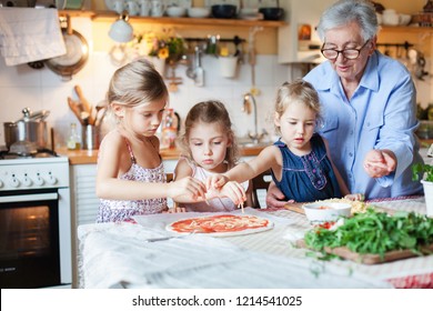 Children And Grandmother Cooking Pizza In Cozy Home Kitchen For Family Dinner. Cute Kids Girls Are Preparing Homemade Italian Food. Three Little Sisters Are Helping Senior Woman. Lifestyle Moment.