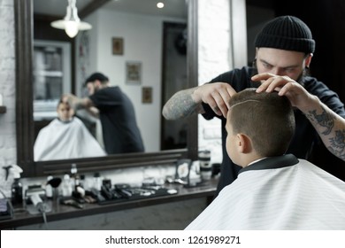 Children hairdresser cutting little boy against a dark background. Contented cute preschooler boy getting the haircut. - Shutterstock ID 1261989271