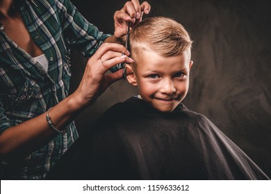 Children hairdresser with scissors is cutting little boy against a dark background. Contented cute preschooler boy getting the haircut. 