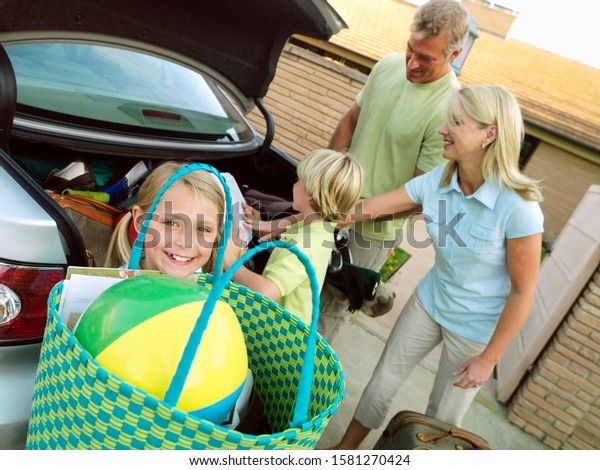 Children help parents load car trunk with\
luggage for family vacation outside\
house