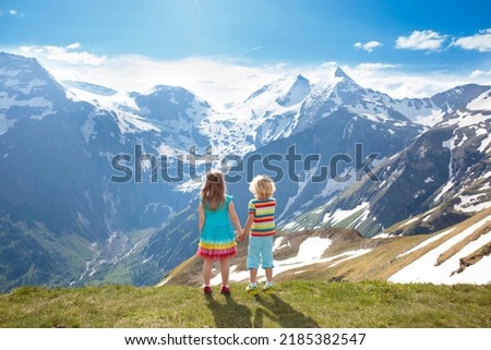 Children hiking in Alps mountains. Kids look at snow covered mountain in Austria. Spring family vacation. Little boy and girl on hike trail in blooming alpine meadow. Outdoor fun and healthy activity.