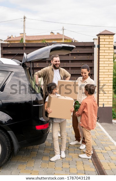 Children holding stuff at car while\
helping parents to unload car during moving into new\
house