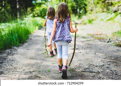 Children - Identical Twins Girls Are Hiking In The Mountains. Active Family, Parents And Children Mountaineering In The Nature. Kids Are Walking In Woods With Walking Sticks.