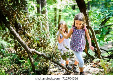 Children - Identical Twins Girls Are Hiking In The Mountains. Active Family, Parents And Children Mountaineering In The Nature. Kids Are Walking In Woods With Walking Sticks.