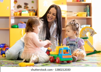 Children Kids Toddlers Playing With Teacher In Kindergarten. Nursery Babies Sitting On Floor Together With Caregiver