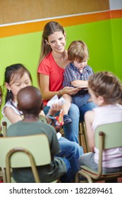 Children In Kindergarten Sitting In Circle And Reading Book With Nursery Teacher