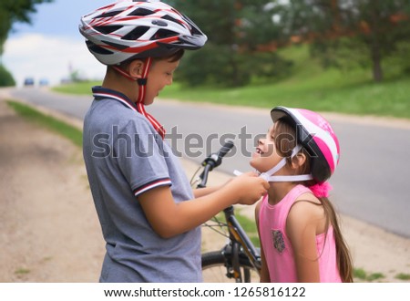 Children learn to ride bicycle in a park on summer day. Teenager boy helping preschooler girl to put on safety helmet. Active outdoor sport for child.