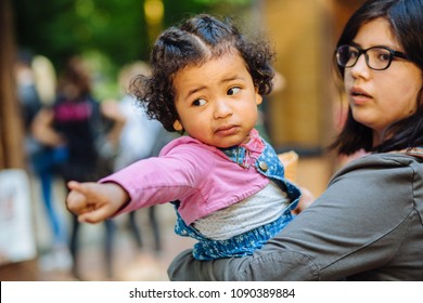 Children Learning And Family Real Life Concept. Candid Moment Of Latino American Mother And Dark Skinned Hispanic Toddler Daughter Crying And Pointing Somewhere During A Walk Outdoors.