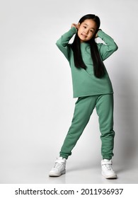 Children, Lifestyle And Body Language. Full Length Asian Girl With Long Hair In A Comfortable Casual Green Suit. Baby Girl Stands On A Light Background And Straightens Her Hair.