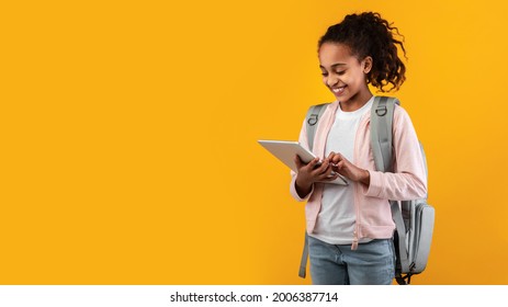 Children And Modern Technologies. Smiling teenage girl wearing backpack holding using digital tablet, studying or playing game standing on yellow orange studio wall, free copy space, panoramic banner - Powered by Shutterstock