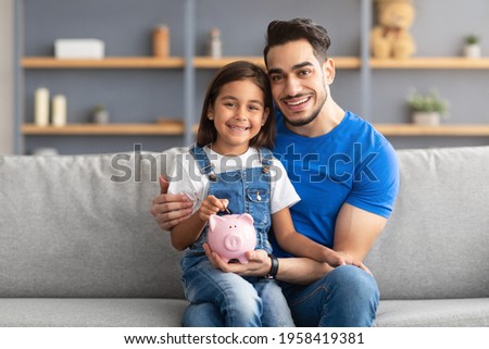 Children and Money Management Cocnept. Portrait of smiling little girl putting coin in pink piggybank, sitting on father's lap and looking at camera, dad teaching daughter to invest for future
