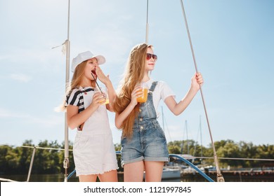 The Children On Board Of Sea Yacht Drinking Orange Juice. The Teen Or Child Girls Against Blue Sky Outdoor. Colorful Clothes. Kids Fashion, Sunny Summer, River And Holidays Concepts.