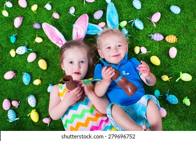 Children On Easter Egg Hunt. Kids Eat Chocolate Rabbit. Boy And Girl Relaxing On A Green Lawn In The Garden. Child Enjoying Sweets. Kid With Bunny Ears.