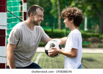 Children And Outdoor Activity Concept. Side view of smiling curly boy holding soccer ball, looking at sportive adult man who tapping and patting son on shoulder, family playing football at pitch