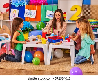 Children painting and drawing in kids club. Art lesson in primary school. Kindergarten teacher who smiling help small students. Kid boy coloring picture on table in class. Balloons on floor.
