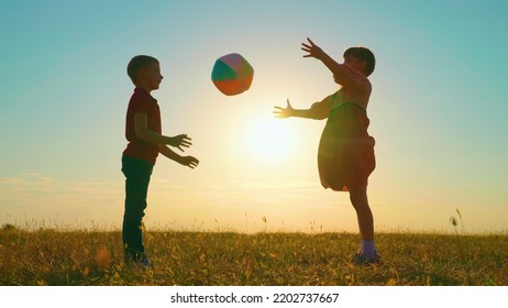 Children Play With Big Ball In Park Against Backdrop Of Sun. Happy Child Throws Ball With His Hands. Kids, Boy, Girl Have Fun With Ball In Park At Sunset. Sports Games For Children Outdoors. Family