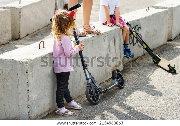 Children play near the concrete blocks. A little\
girl stands next to a scooter and a concrete fence. Close-up.\
unrecognizable person
