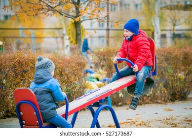 Children Play On The Playground Outside. Group Of Children Playing Together In Kindergarten Outside. Happy Excited Kids Having Fun Together On Playground. Children Ride On A Swing
