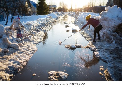 Children Play In Puddle On Street In Early Spring. Snow Melting On Road. Weather Thaw. Fun Game With Boat And Water Outdoors In Countryside.