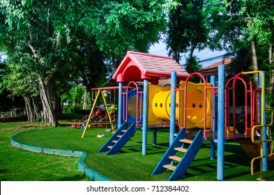 Children playground on yard activities in public park surrounded by green trees at sunny morning day. Children run, slide, swing,seesaw on modern playground. Urban neighborhood childhood concept.