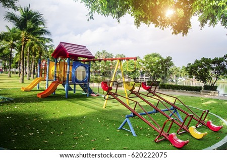  Children playground on yard activities in public park surrounded by green trees at sunlight morning. Children run, slide, swing,seesaw on modern playground. Urban neighborhood childhood concept.