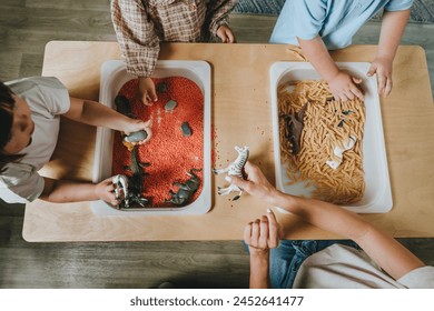 Children playing and developing tactility with toy animal figures in bulk cereals and pasta with teacher in kindergarten. Selective focus, top view.