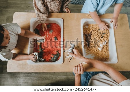 Children playing and developing tactility with toy animal figures in bulk cereals and pasta with teacher in kindergarten. Selective focus, top view.