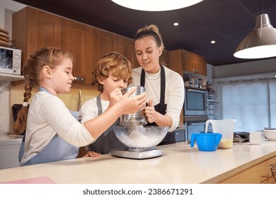 Children pouring flour from sieve in bowl on scales while helping mom in kitchen and enjoying cooking together