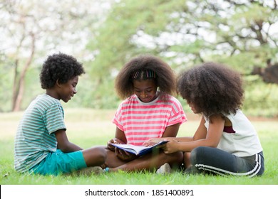 Children reading book on grass outdoor. Group of African American children in casual wear reading book while sitting on green grass in the park, against green summer garden