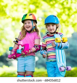 Children Riding Skateboard In Summer Park. Little Girl And Boy Learn To Ride Skate Board, Help And Support Each Other. Active Outdoor Sport For Kids. Child Skateboarding. Preschooler Kid Skating.