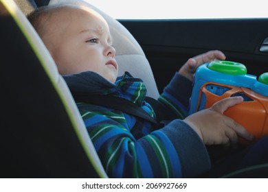 Children Road Safety Concept. Upset Toddler Boy Traveling In Car Sitting In Child Safety Seat With Toy Car.