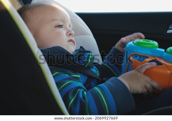 Children road safety\
concept. Upset toddler boy traveling in car sitting in child safety\
seat with toy car.
