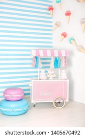 Children Room With Blue Stripe Background. Candy Stall Photo Zone With Big Macaroons, Sweets And Marshmallows. Trolley With Ice Cream.  Decorated Room For Birthday. Cart With Candy Bar. Birtday Decor