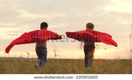 Children running with red cloaks on backs blowing in wind at sunset across field. Children running freely play favourite superheroes at sunset on field. Children dressed as superheroes run to sunset