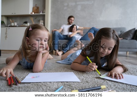 Children sisters playing drawing together on floor while young parents relaxing at home on sofa, little girls having fun