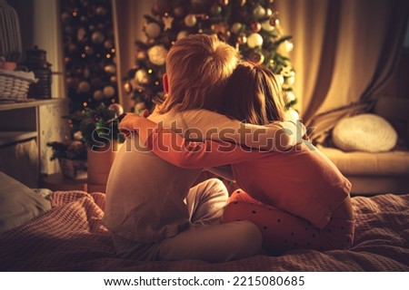 Children sit hugging in bed and look at the Christmas tree. Christmas decorations, waiting for the holiday. 