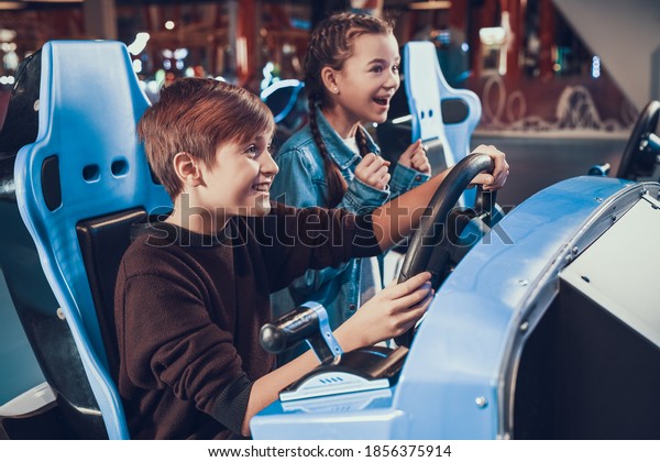 Children\
sit at slot machines and have fun. Cheerful sister cheering on\
brother who drives a toy car in a video game.\
